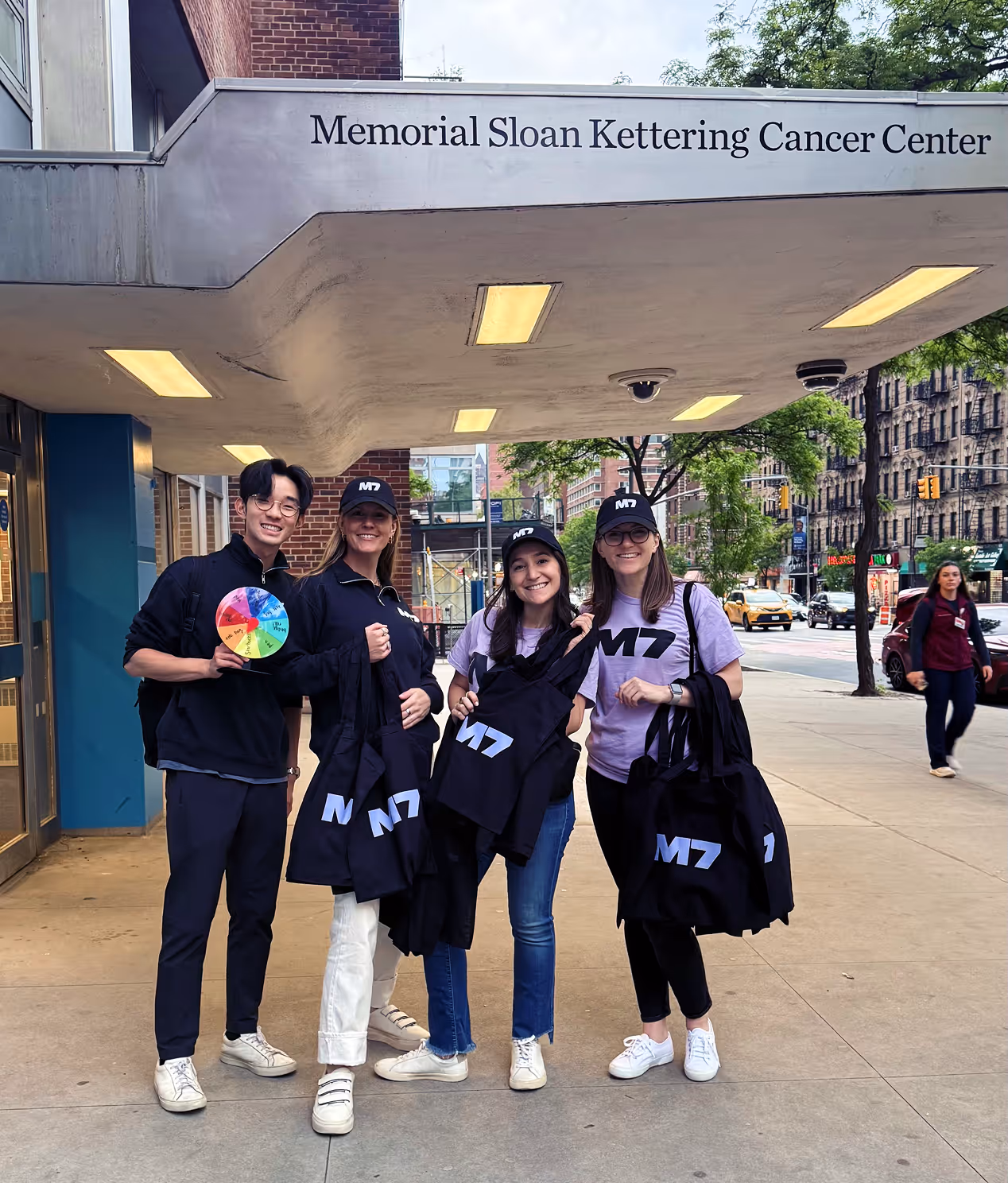 Four smiling young adults holding black tote bags with M7 logo, standing under Memorial Sloan Kettering Cancer Center entrance.