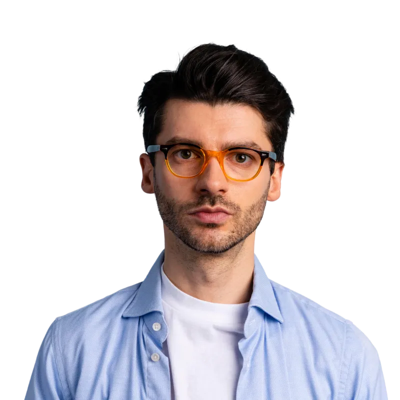 Young man with black hair and light beard wearing amber-framed glasses and a blue shirt over a white t-shirt.