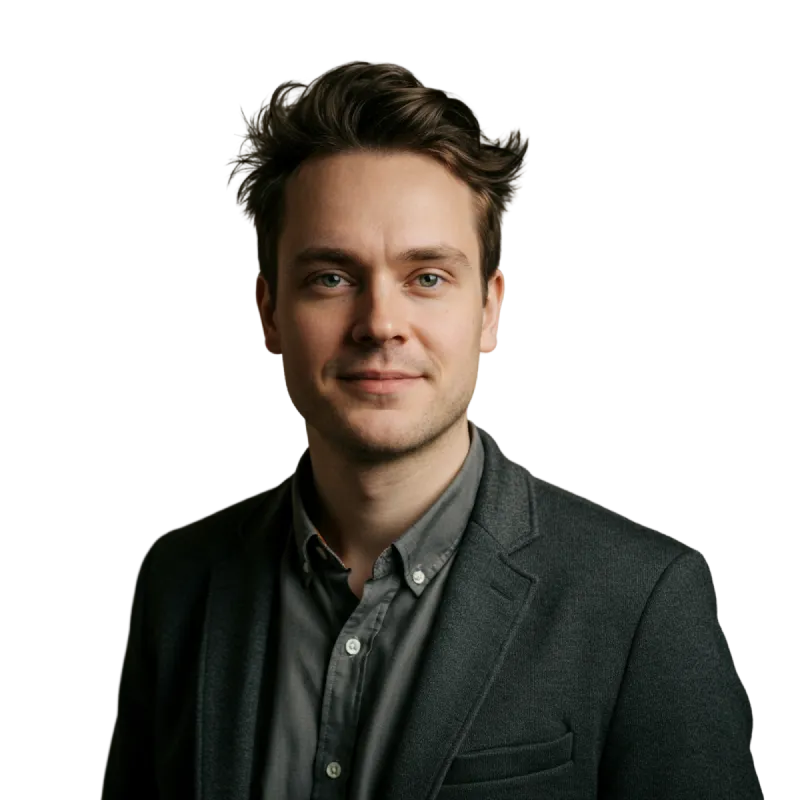 Portrait of a man with light skin and styled brown hair wearing a dark gray blazer and buttoned shirt, looking at the camera with a slight smile.