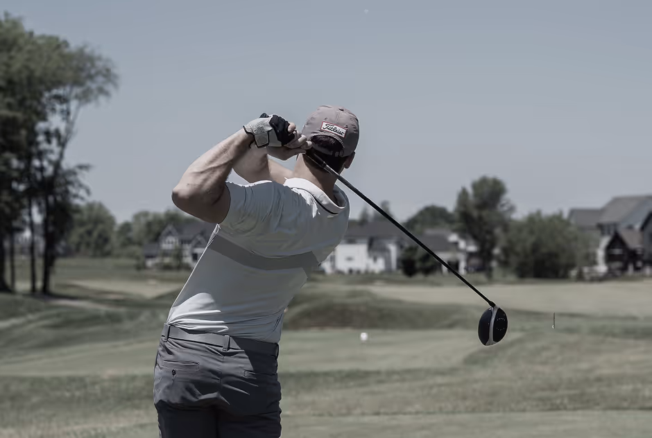 Golfer in mid-swing driving a golf ball on a course with houses and trees in the background.