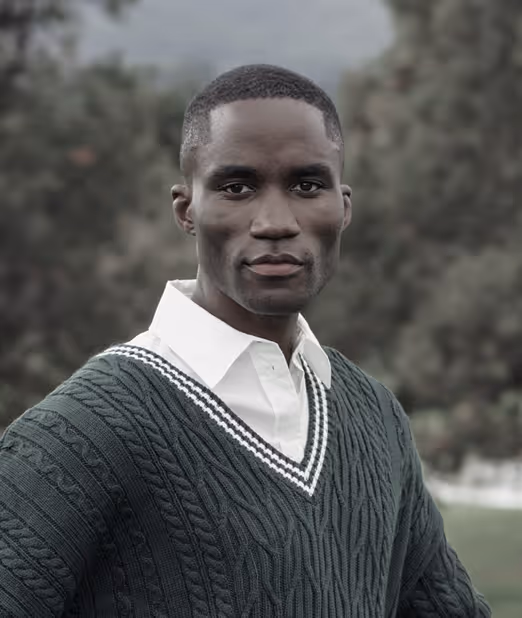 Portrait of a serious young man wearing a green cable-knit sweater over a white collared shirt outdoors with blurred trees in the background.