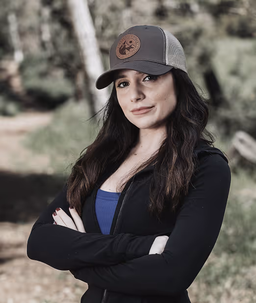Young woman with long dark hair wearing a baseball cap and black jacket standing outdoors with arms crossed.