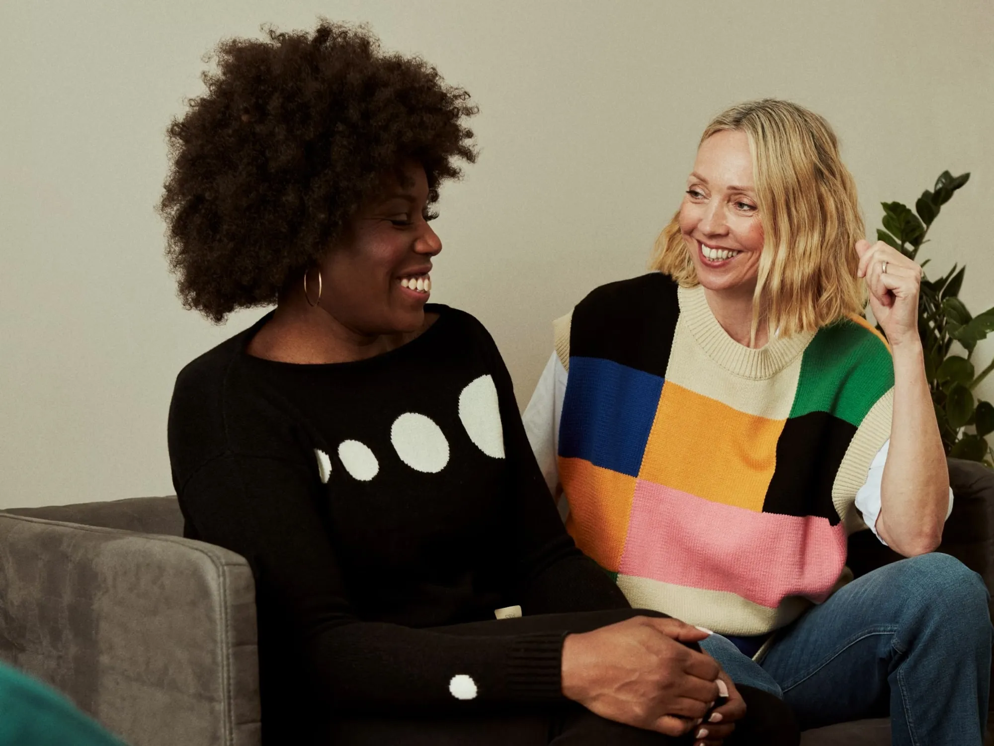 Zwei Frauen sitzen auf einem Sofa und lachen miteinander, eine trägt einen schwarzen Pullover mit weißen Punkten, die andere einen bunten, karierten Pullover.