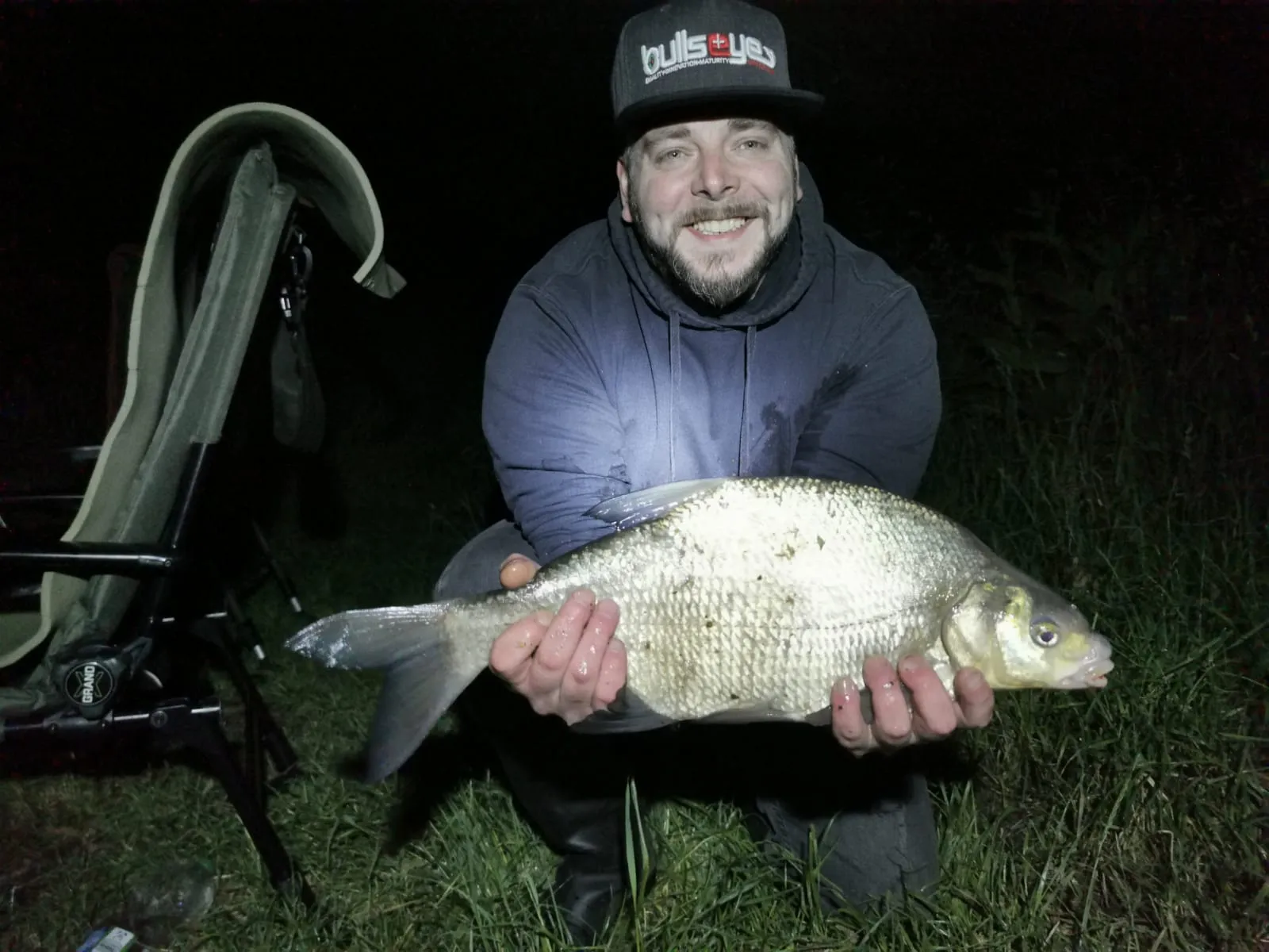 Mann kniet im Gras und hält einen großen silbernen Fisch bei Nacht, ein Angelstuhl steht links.