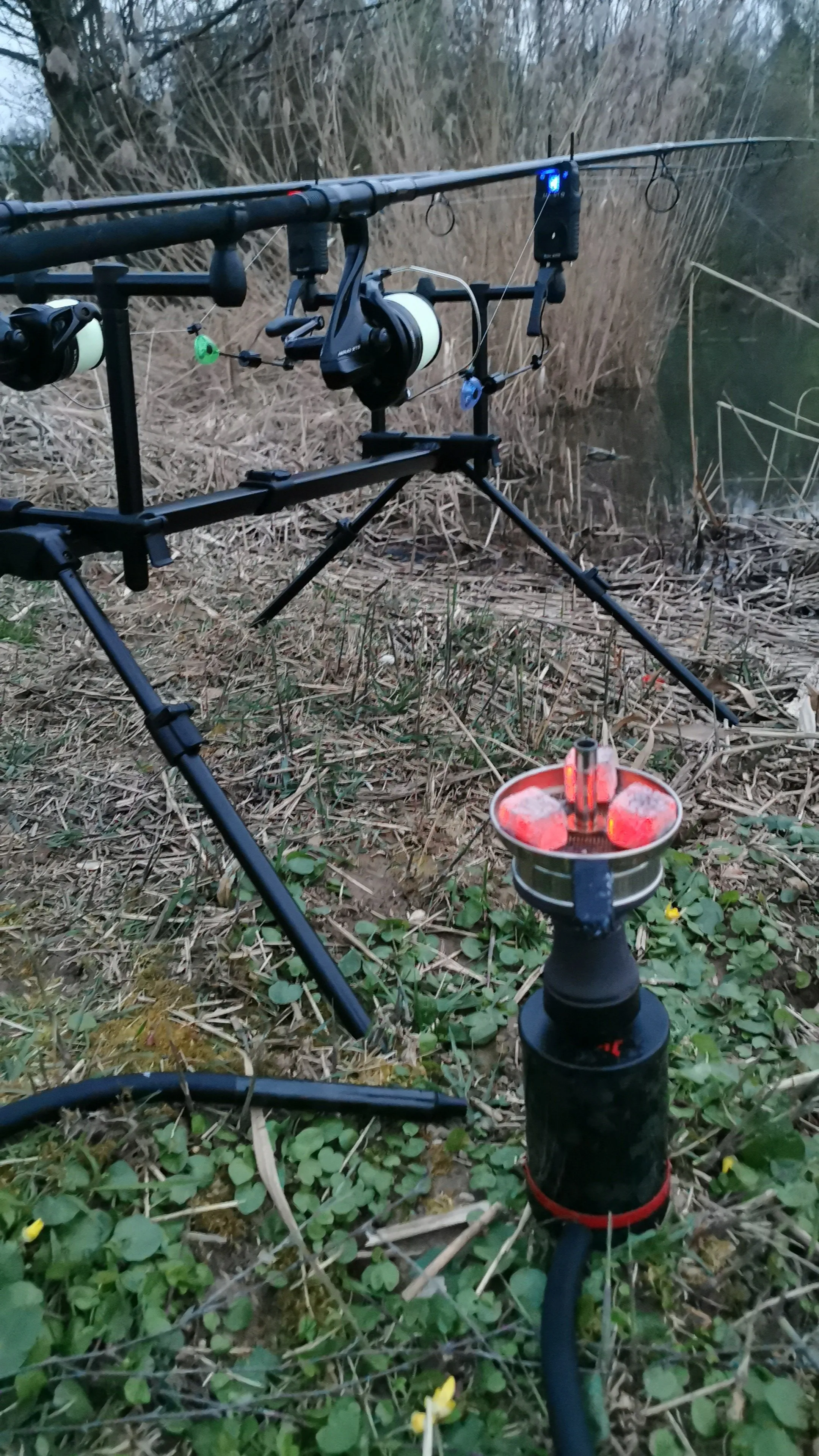 Angelruten auf einer Halterung am Ufer eines Teichs mit einem brennenden Köderfeuer im Vordergrund.