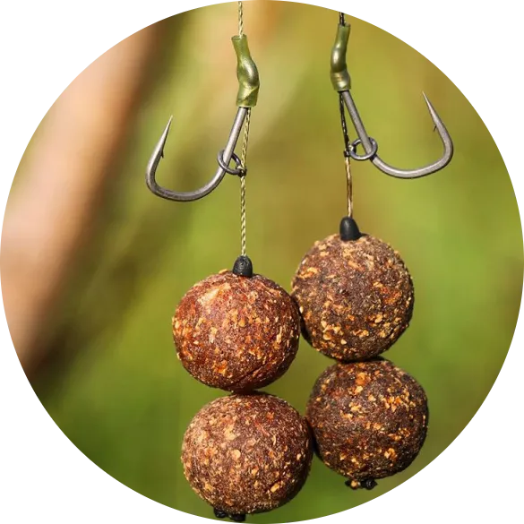 Zwei Angelhaken mit jeweils zwei braunen Köderkugeln aus körnigem Material an einer Angelschnur, vor unscharf grünem Hintergrund.