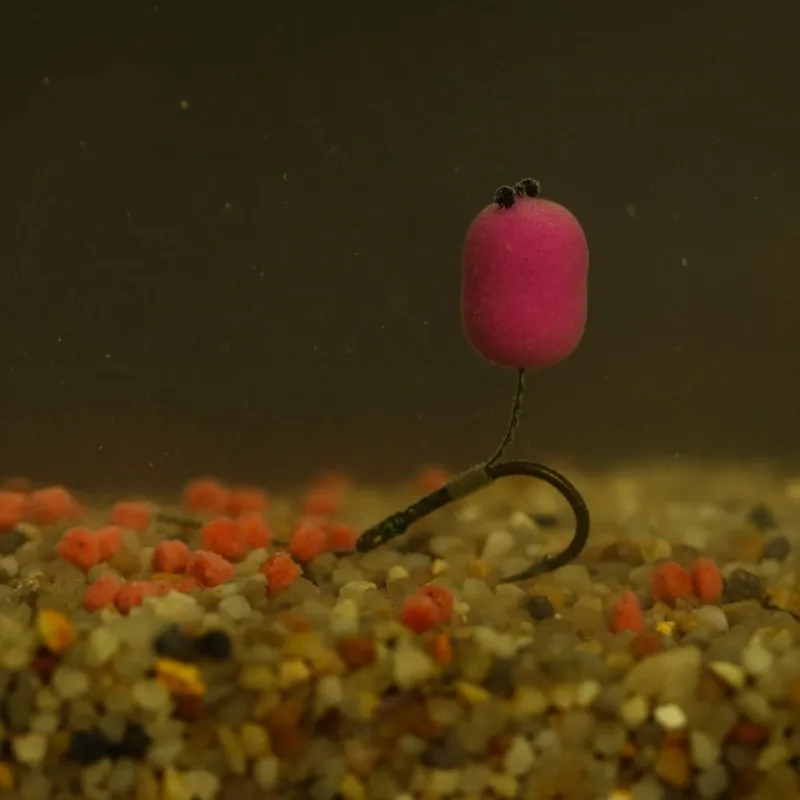 Unter Wasser platzierter Angelhaken mit einem pinken Köder und kleinen roten Partikeln am Kiesboden.