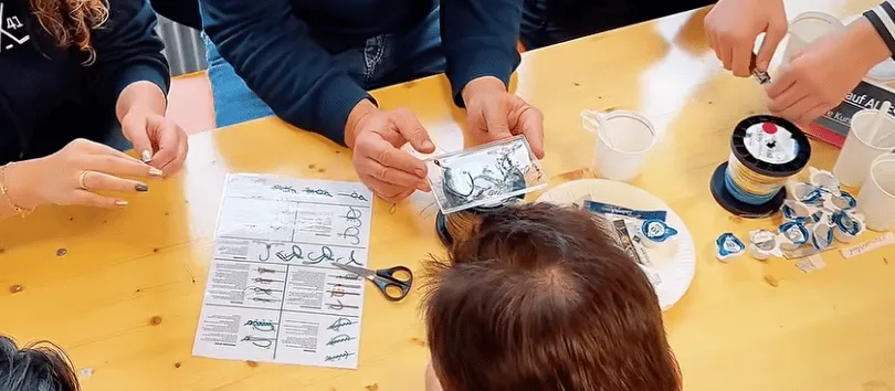 Mehrere Personen sitzen um einen Holztisch und halten Angelhaken, Angelsehnen und einen Anleitungszettel in der Hand.