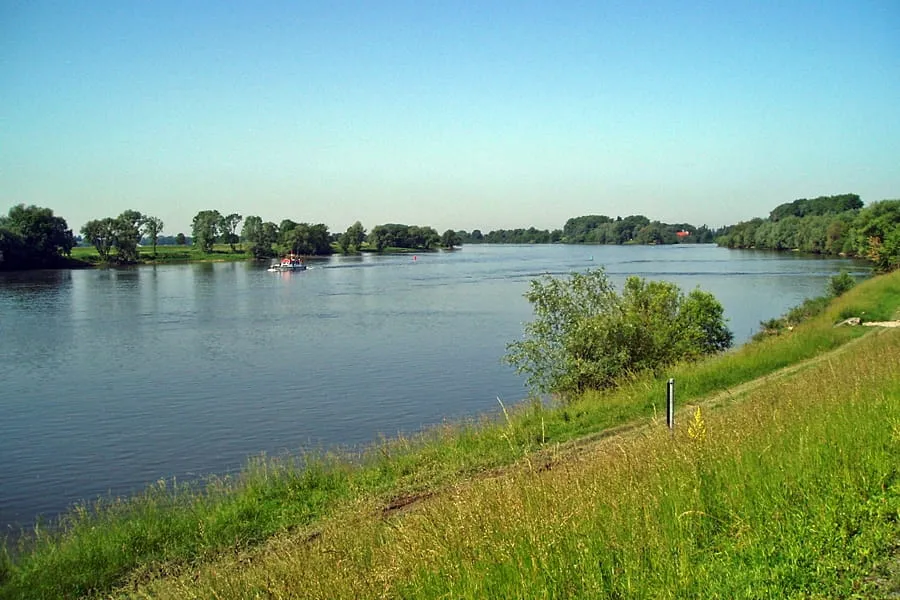 Breiter Fluss mit grün bewachsenen Uferbereichen und einem Boot auf dem Wasser unter klarem Himmel.