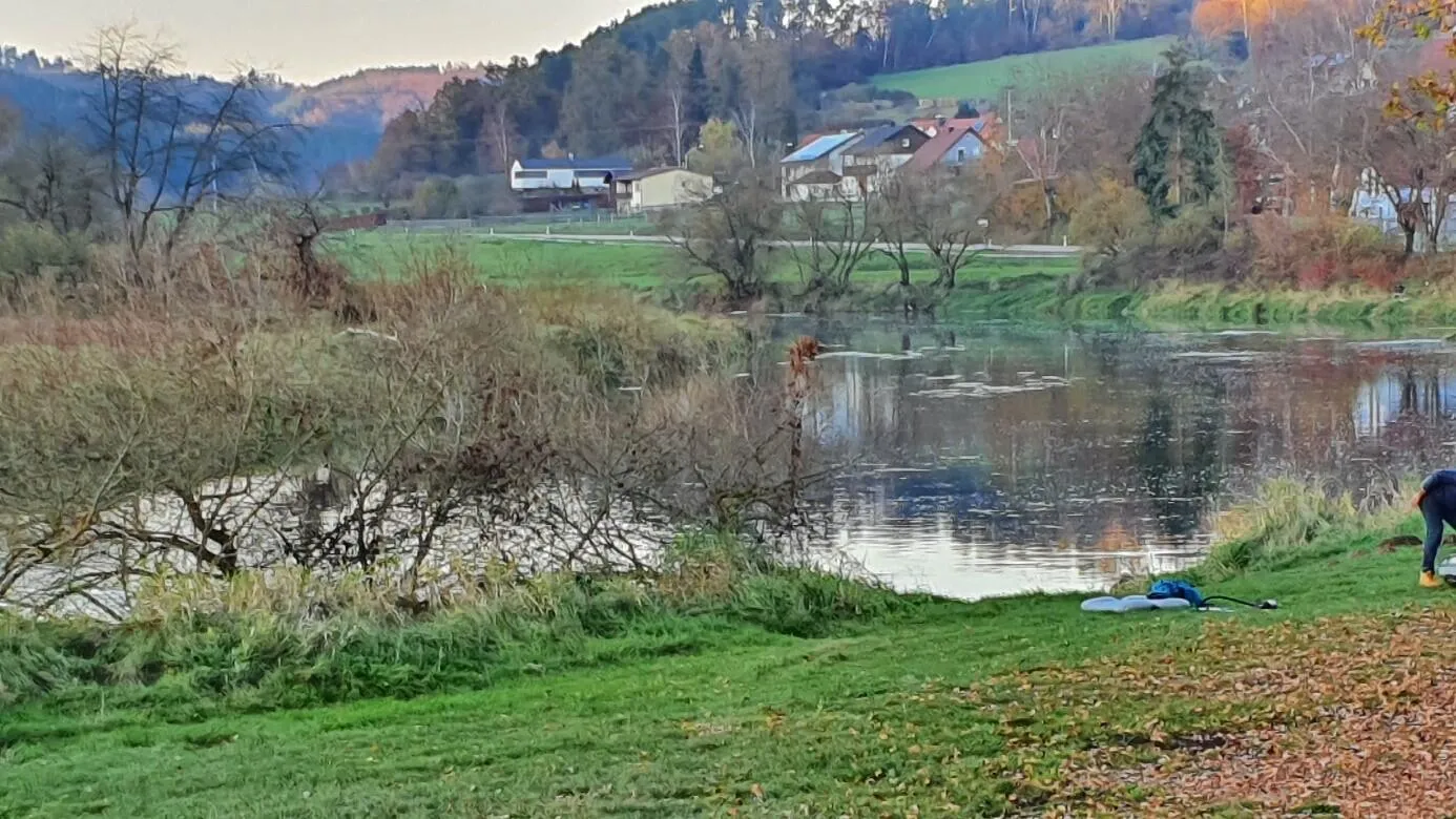 Flussufer mit grünem Gras, Bäumen, einigen Häusern im Hintergrund und einer Person, die sich am Wasser bückt.