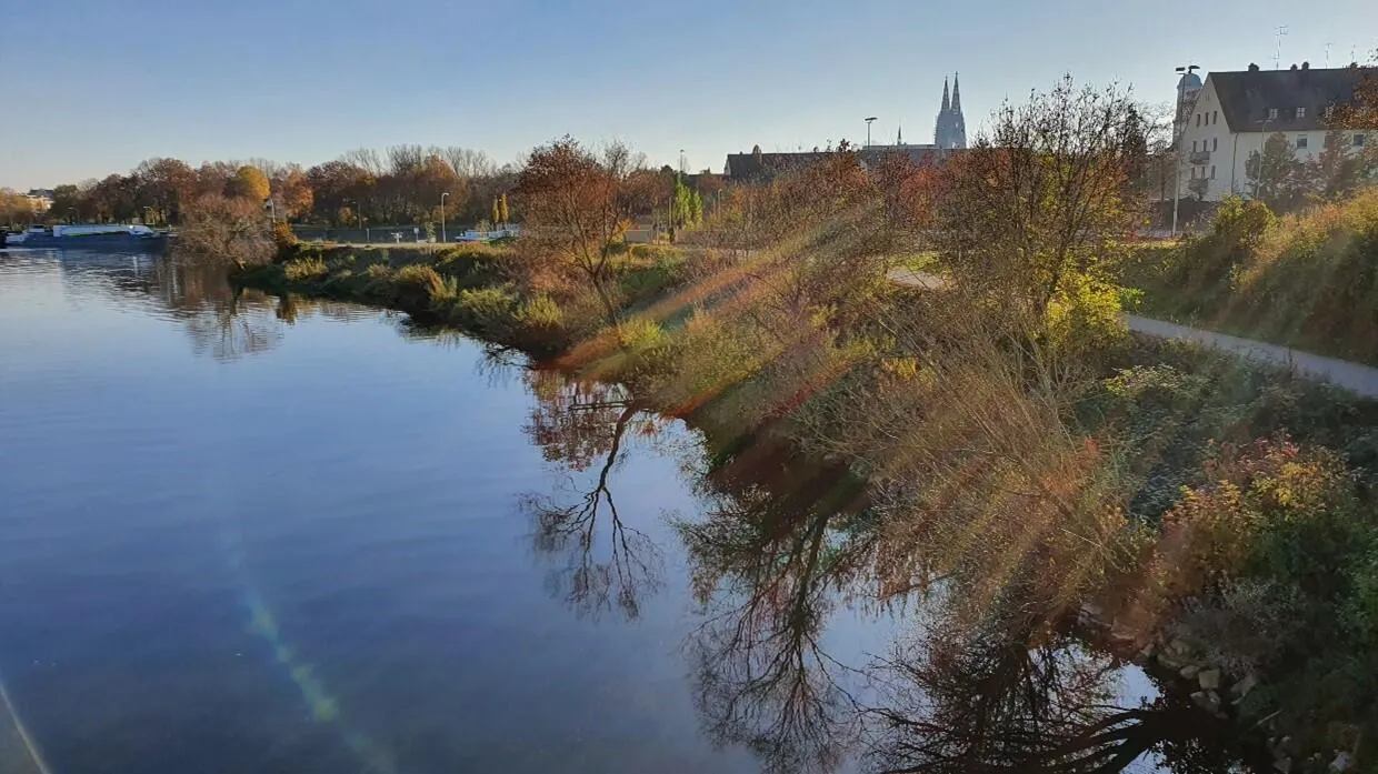 Herbstliche Flusslandschaft mit reflektierten Bäumen im ruhigen Wasser und Stadtkulisse mit Domtürmen im Hintergrund.