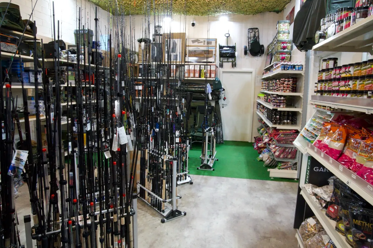 Angelgeschäft mit zahlreichen Angelruten in der Mitte, Regalen mit Angelzubehör und Ködern an den Seiten und grünem Teppich im hinteren Bereich.