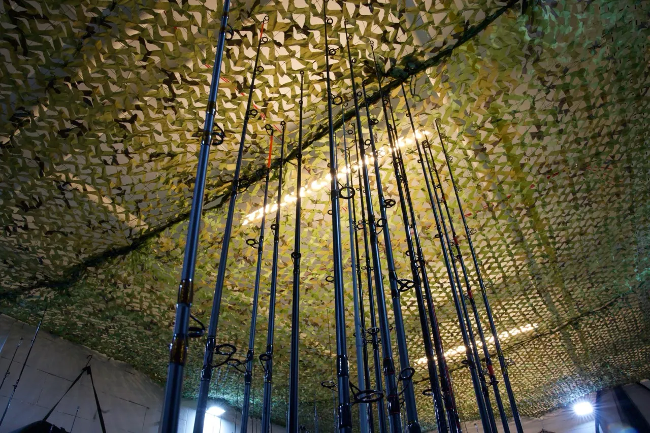 Mehrere vertikal stehende Angelruten vor einer Decke mit camouflage Tarnnetz in einem Innenraum.