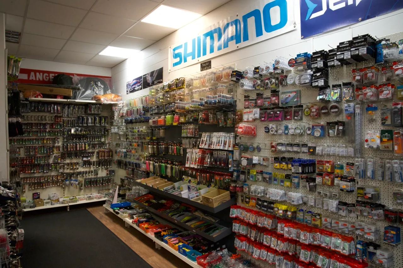 Innenansicht eines Angelgeschäfts mit einer großen Auswahl an Angelschnüren, Ködern und Angelzubehör an den Wänden, darunter ein großes Shimano-Banner.