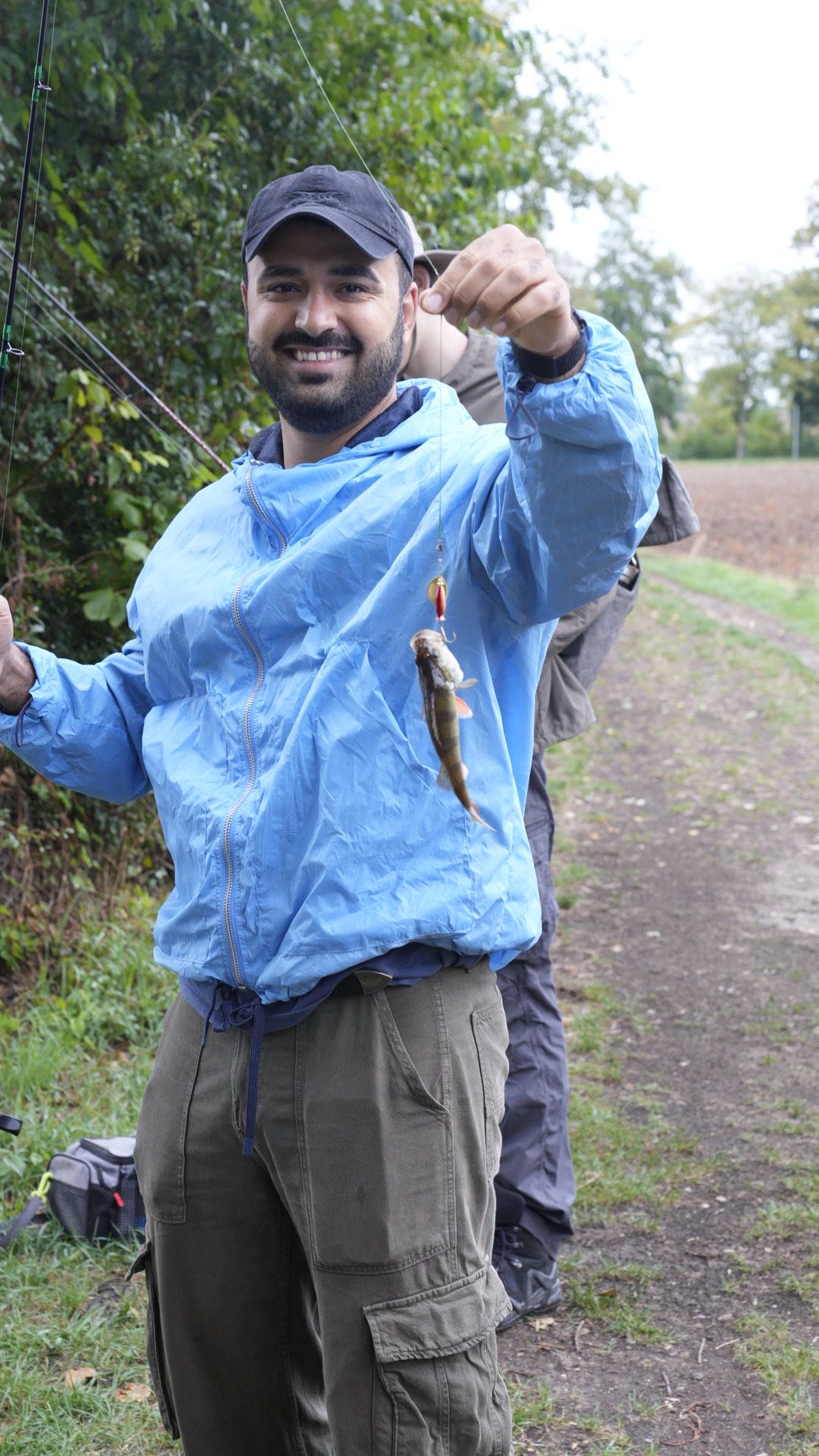 Lächelnder Mann mit blauer Jacke und schwarzer Mütze hält eine Angelrute und zeigt einen kleinen Fisch, den er gefangen hat; im Hintergrund sind Bäume und ein Weg sichtbar.