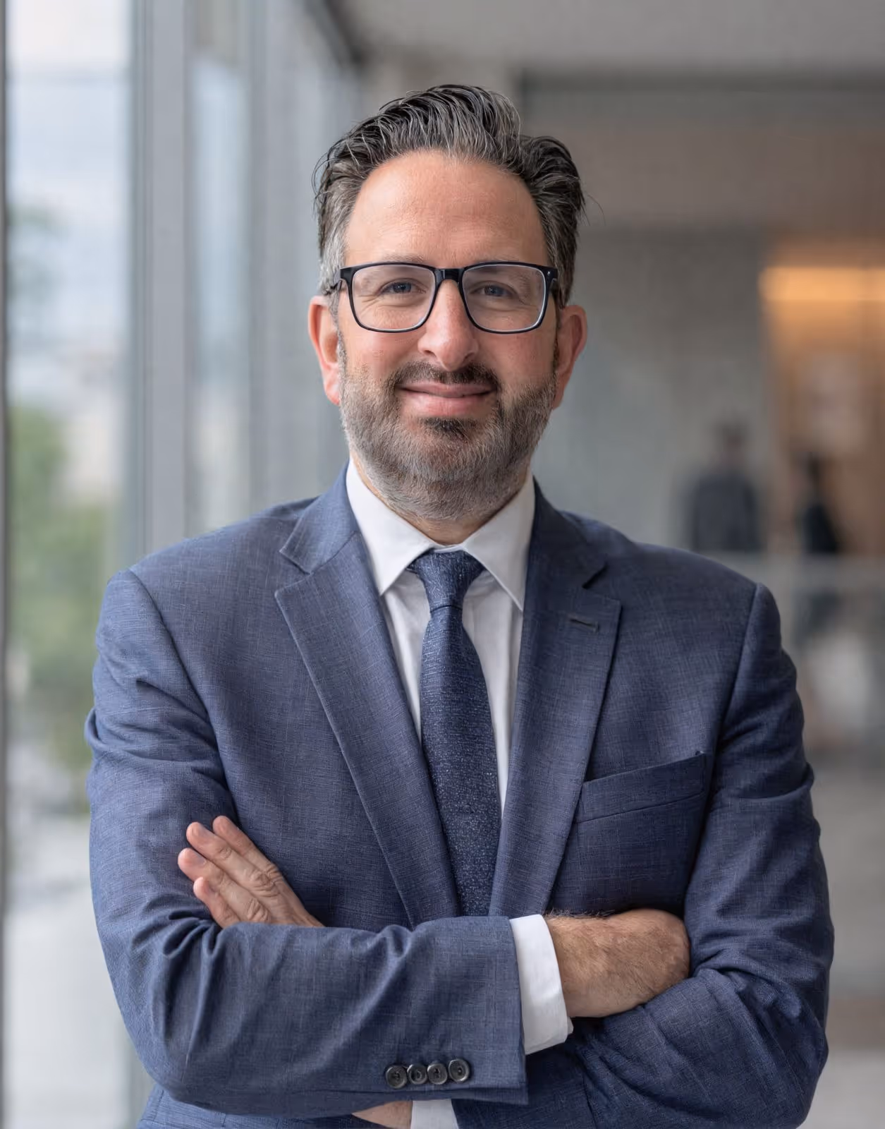Man with glasses and beard wearing a blue suit and tie, standing indoors with arms crossed and smiling.