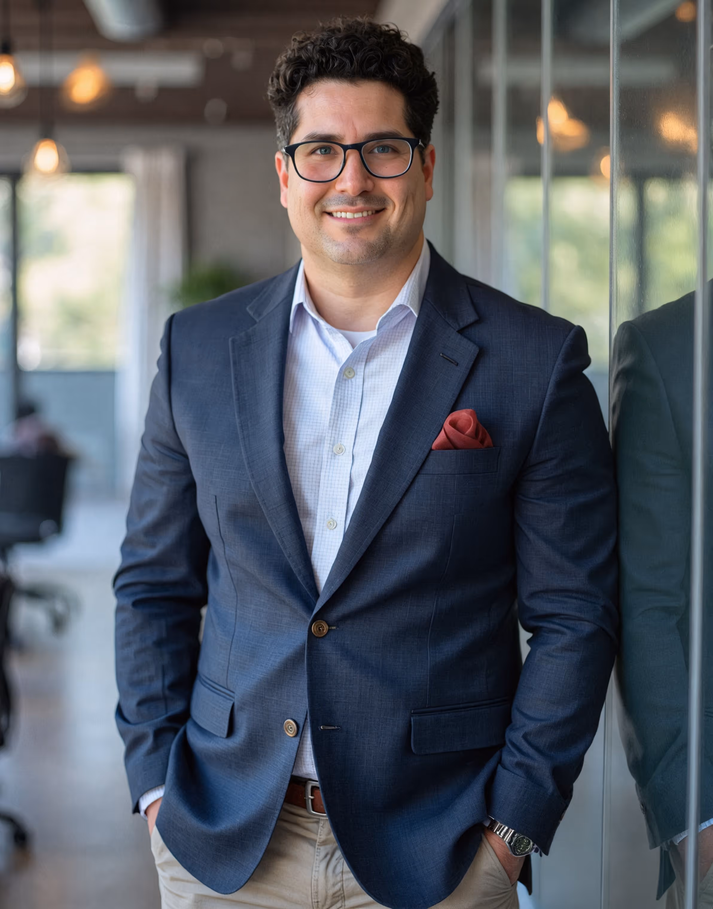 Man with curly dark hair and glasses wearing a navy blazer with a red pocket square, standing with hands in pockets next to a glass wall in an office.