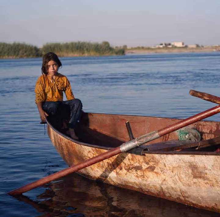 Young girl wearing a yellow shirt sitting at the edge of a rusty wooden boat on calm water during daytime.