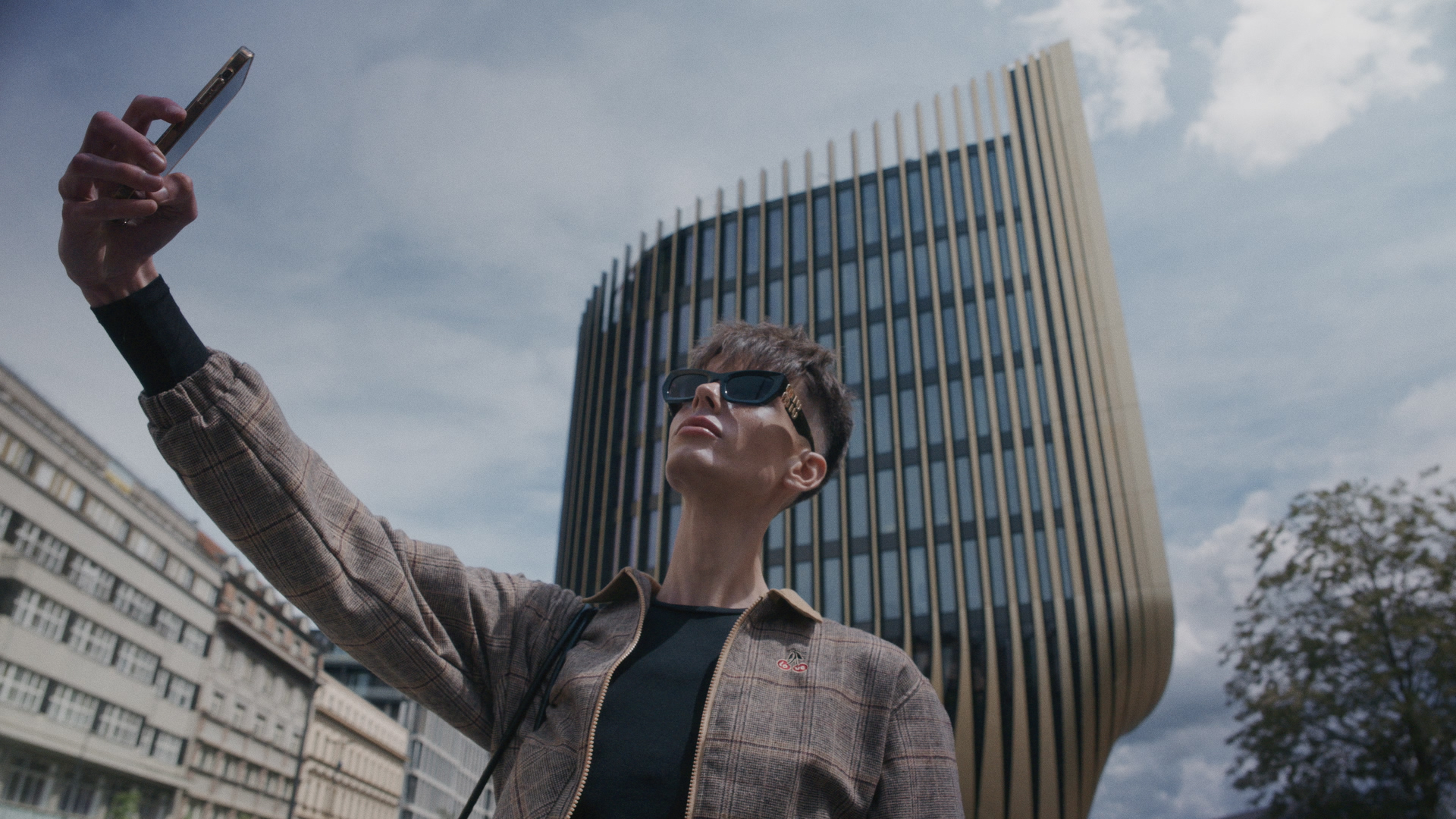 Person wearing sunglasses and a plaid jacket taking a selfie outdoors with modern buildings in background.