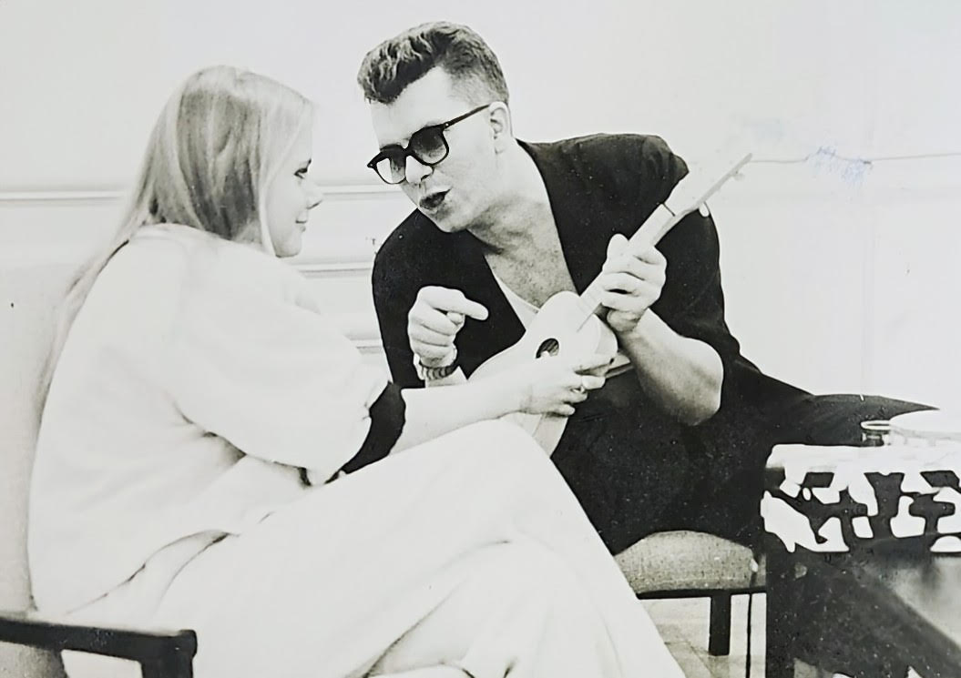 A man shows a woman how to play a small guitar-like instrument while seated indoors.
