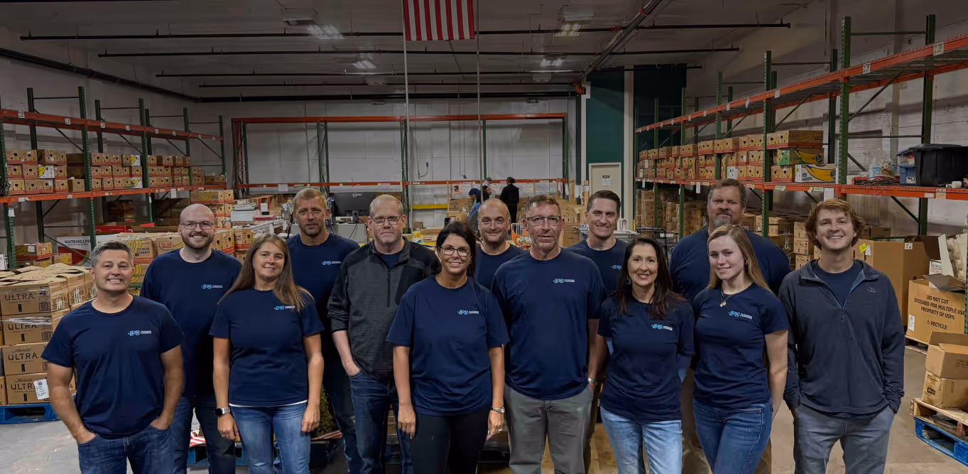 A group of 13 diverse people standing together and smiling inside a warehouse filled with shelves and boxes, with an American flag hanging from the ceiling.