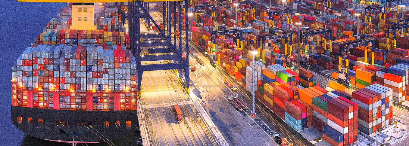 Large cargo ship loaded with stacked shipping containers docked at a brightly lit port full of colorful containers and cranes at night.
