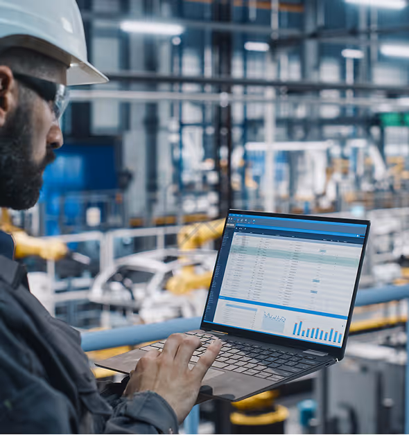 Engineer wearing a white hard hat and glasses working on a laptop with data analytics displayed, inside an industrial factory with robotic arms.
