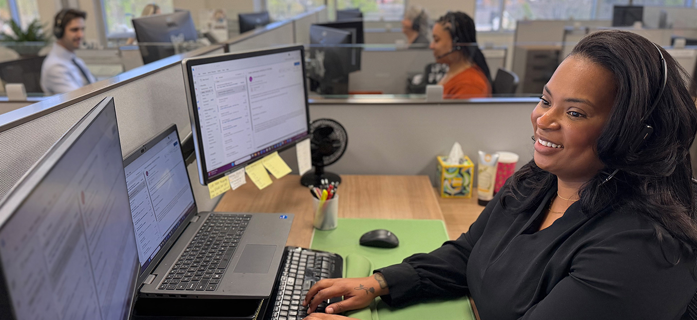 Smiling woman with headset working at a computer in an office call center with colleagues in the background.