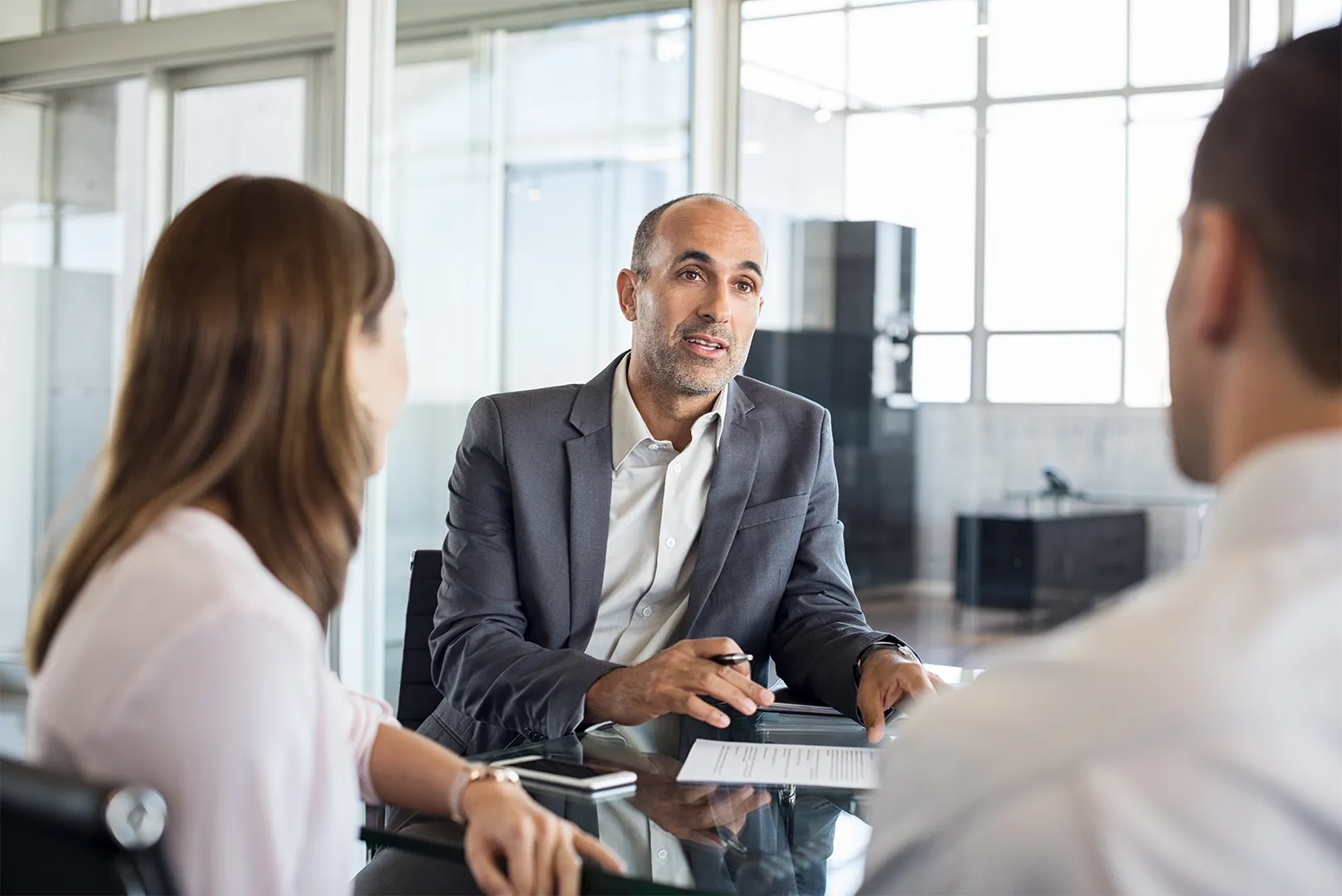Businessman in a suit talking to a couple across a glass table in a modern office.