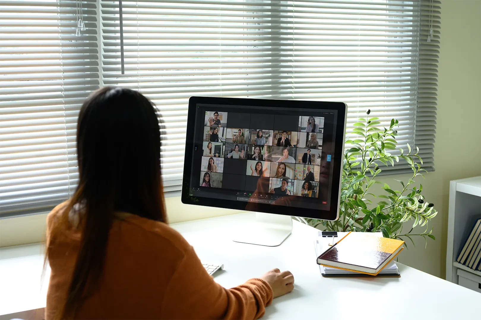 Person with long hair in an orange sweater attending a video conference on a desktop computer in a bright office with blinds and a green plant nearby.