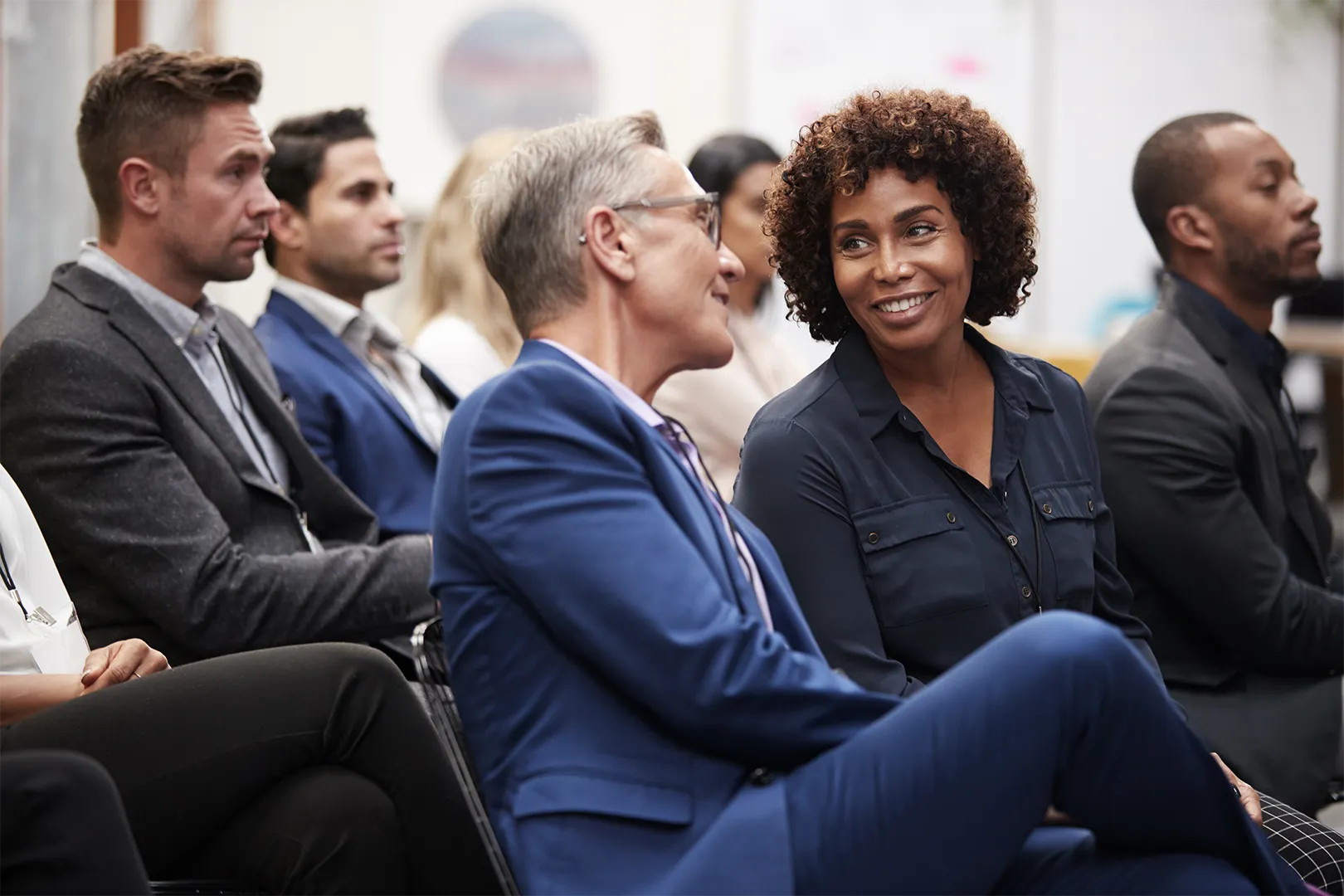 A diverse group of professionals seated in a conference room, with two individuals in the foreground engaged in a friendly conversation.