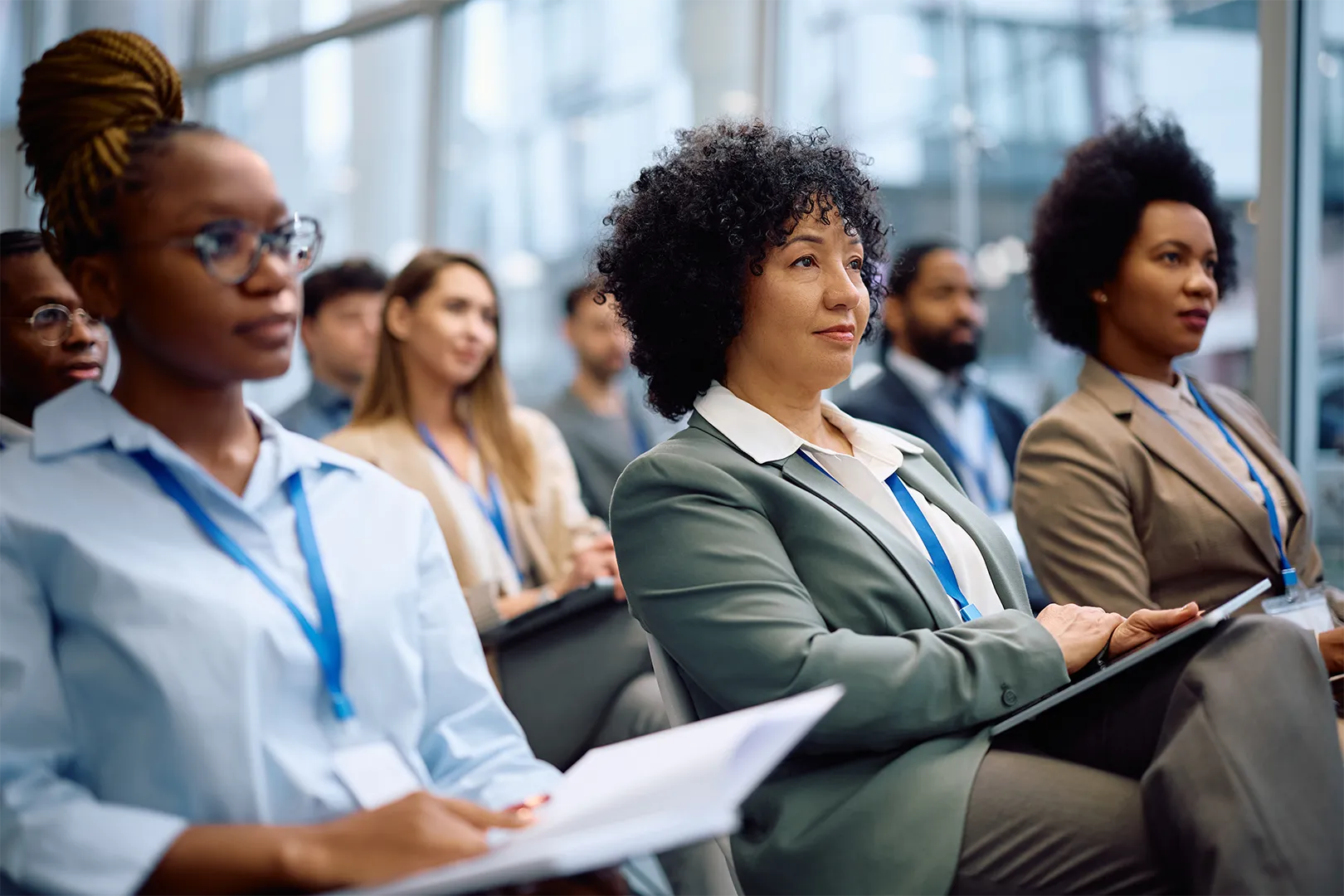 Diverse group of professionals attentively listening at a conference, some holding notebooks or documents.