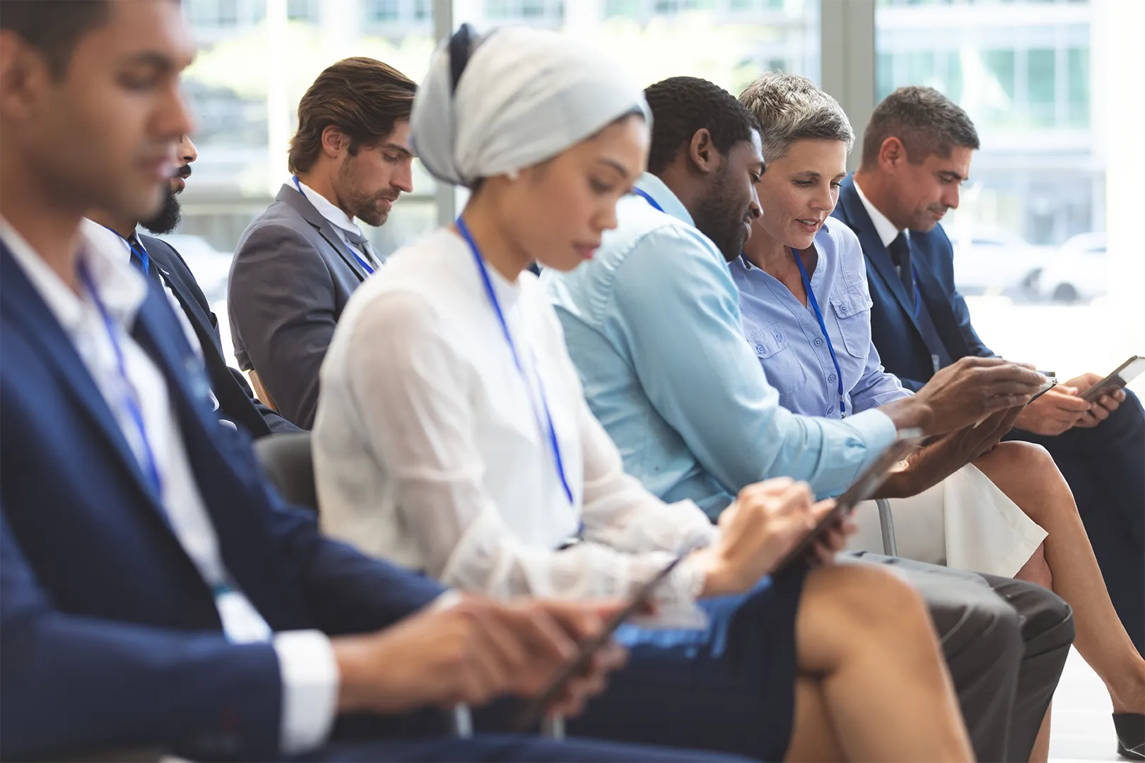 Diverse group of business professionals sitting in a row, focused on tablets or smartphones during a conference.