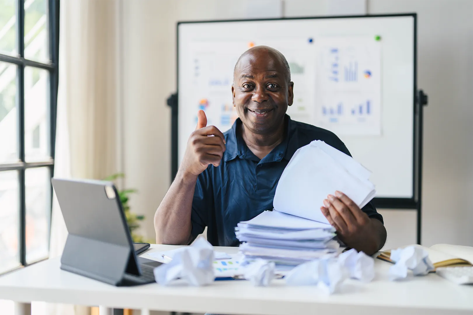 Smiling man sitting at a desk with a large stack of papers, giving a thumbs-up gesture.