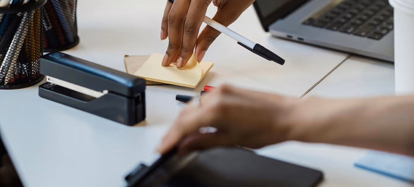 Two hands working on a desk with a pen, sticky notes, a stapler, and a laptop nearby.