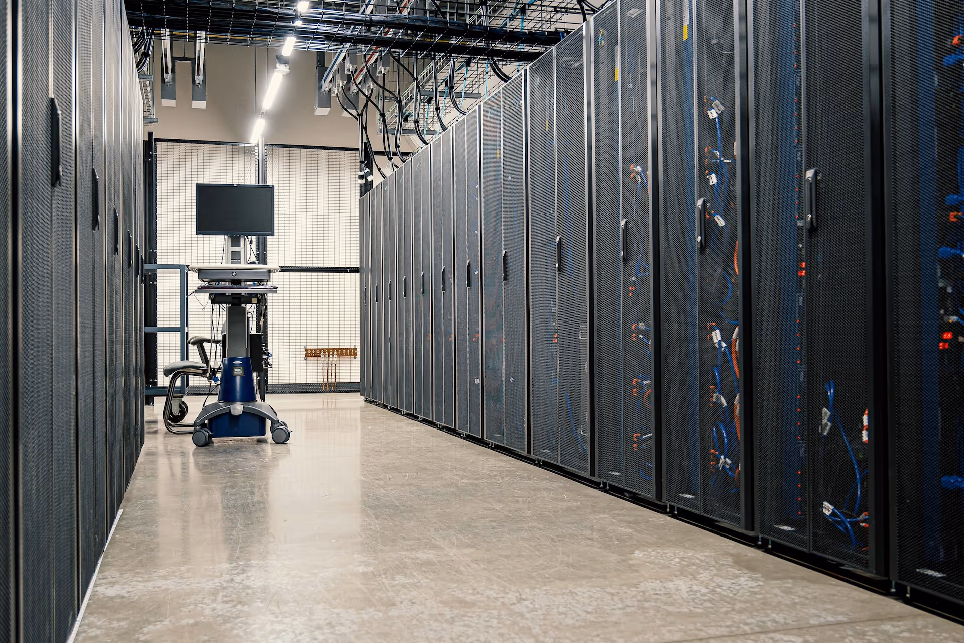 Interior view of a server room with rows of black server racks and a mobile computer workstation with a monitor and keyboard in the center.