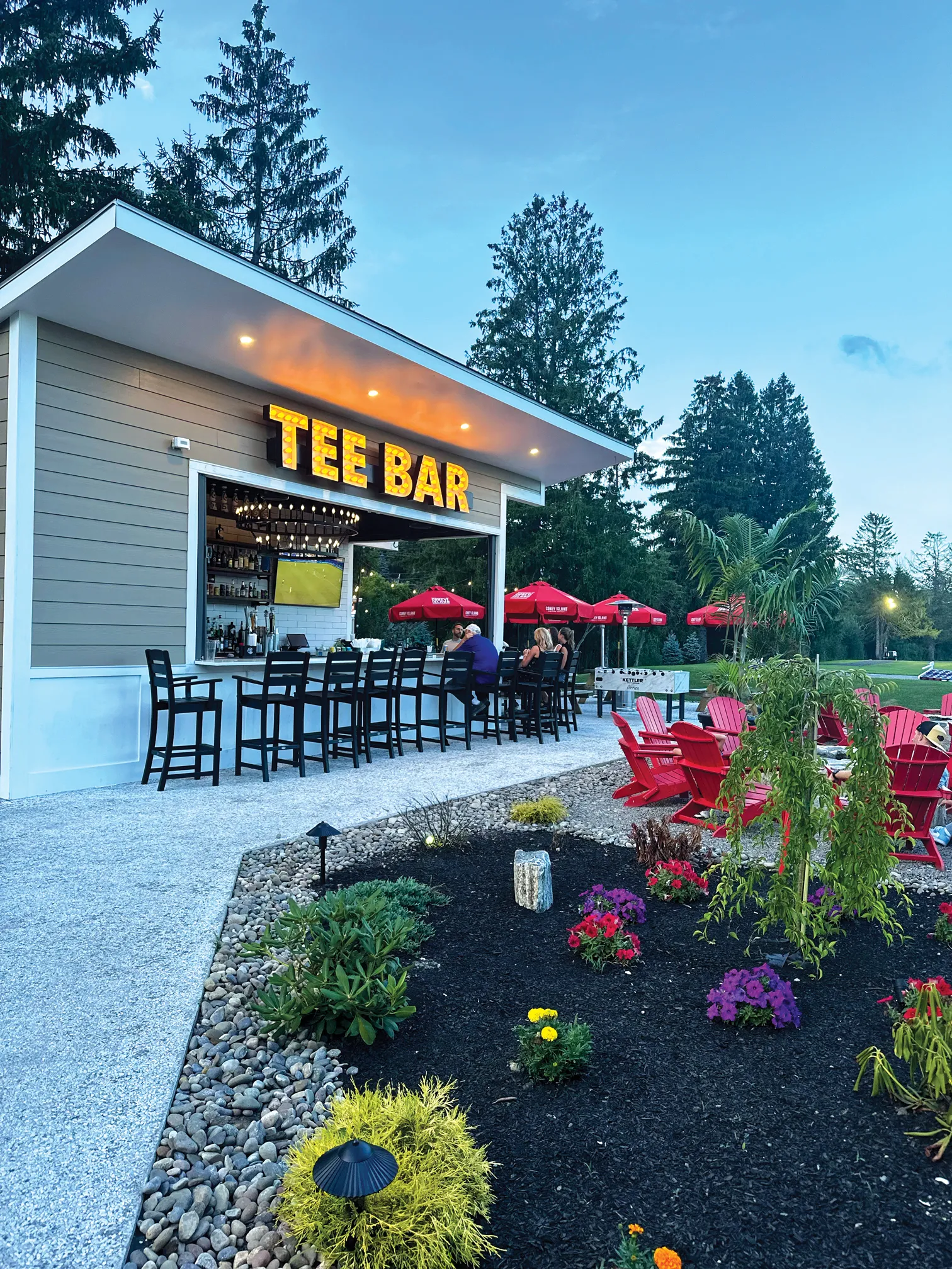 Outdoor bar named Tee Bar with black chairs along the counter and red umbrellas in the seating area surrounded by greenery and flowers during dusk.