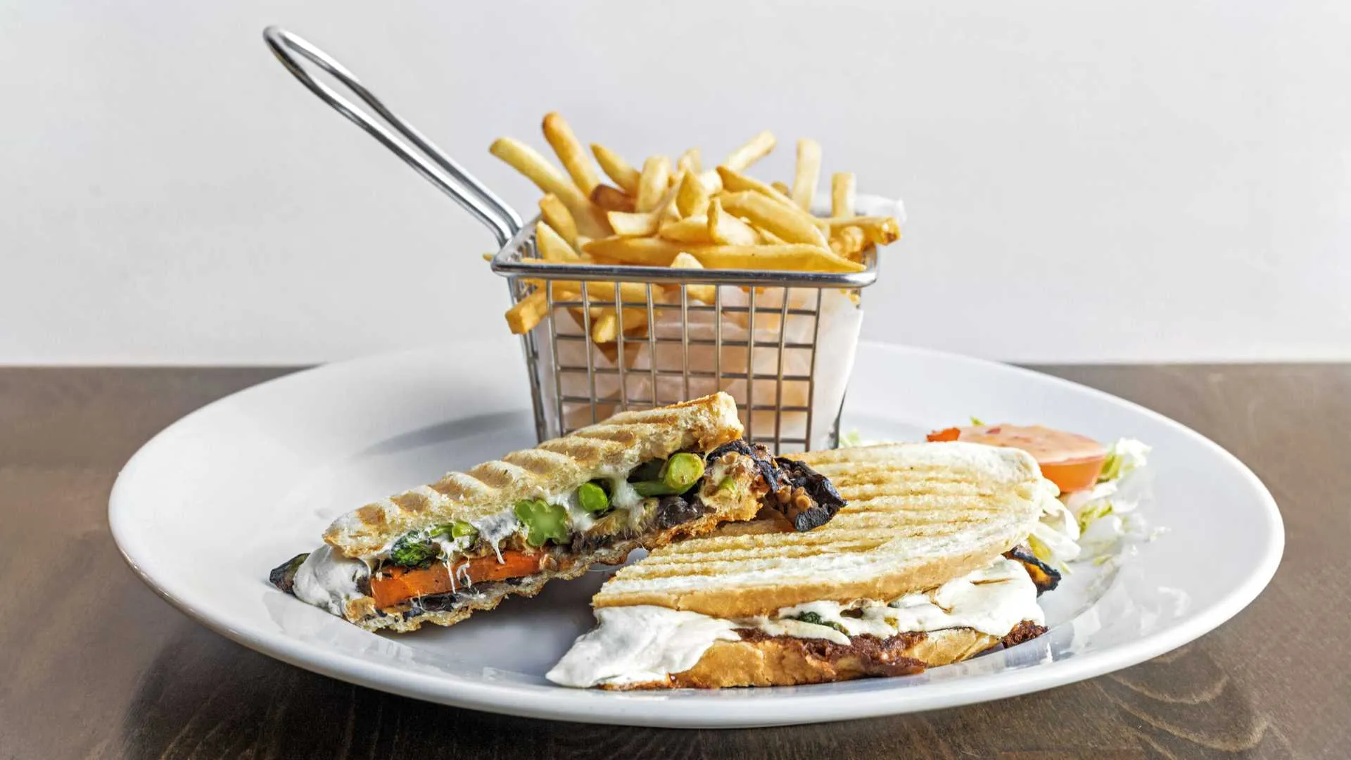 Grilled vegetable panini sandwich with melted cheese and asparagus, served with french fries in a small metal basket on a white plate.