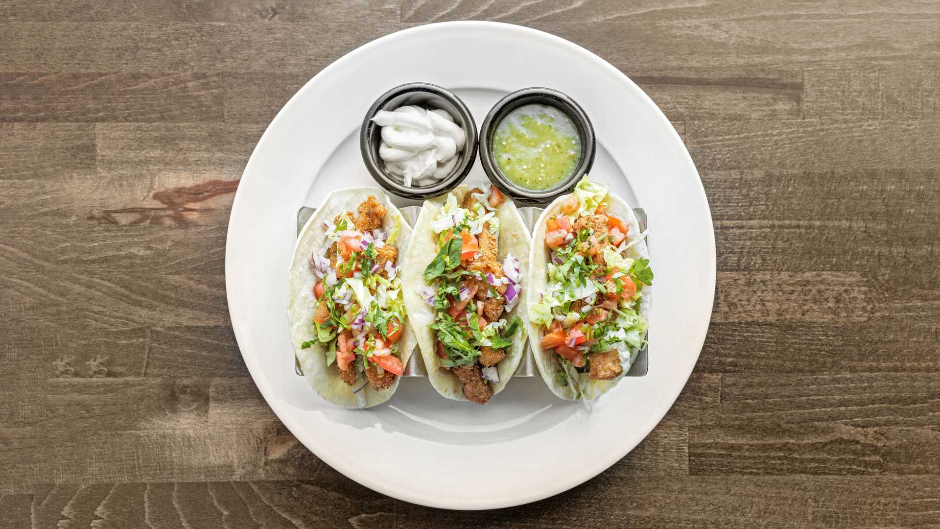 Three fish tacos with shredded lettuce, diced tomatoes, onions, and cilantro on a white plate with cups of sour cream and green salsa.