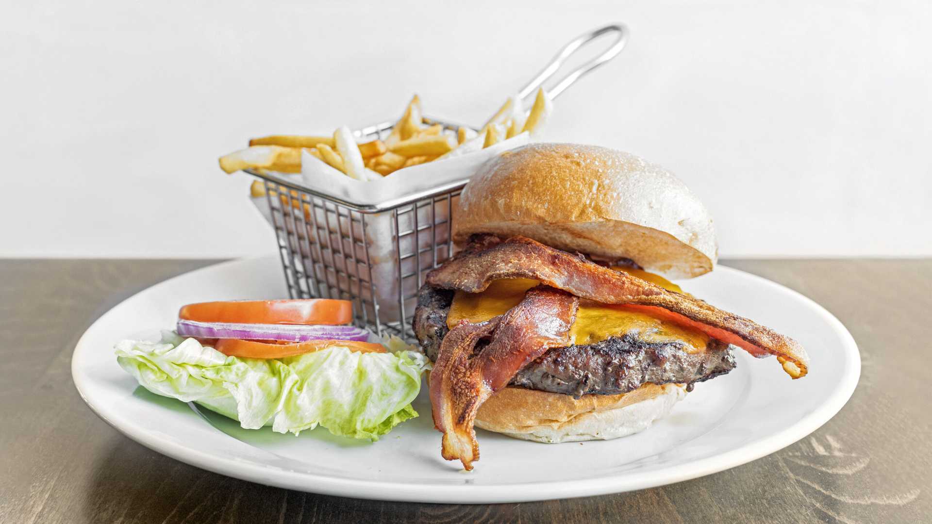 Bacon cheeseburger with lettuce, tomato, and onion on a white plate served with a basket of French fries.