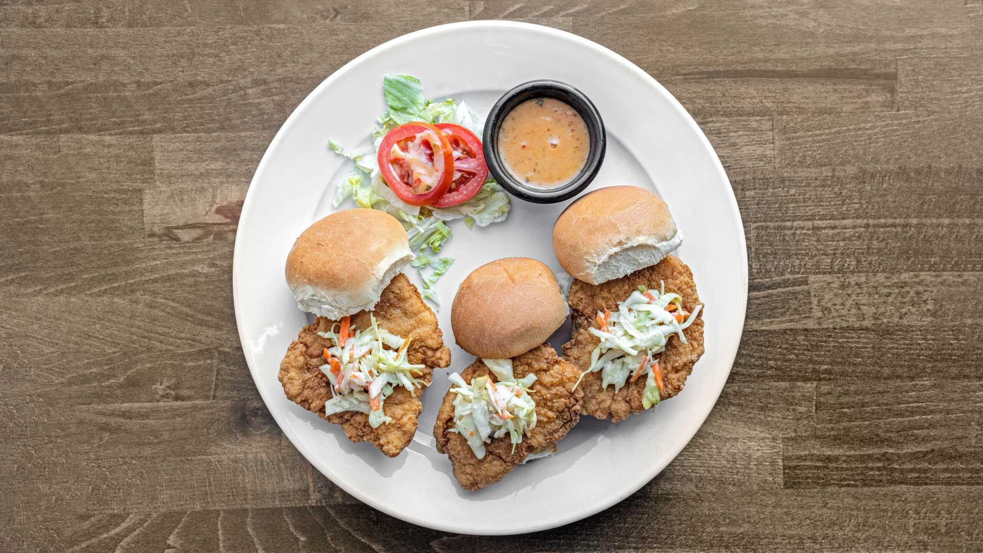 Plate with three fried chicken sliders topped with coleslaw, a small cup of dipping sauce, and sliced tomatoes on shredded lettuce.
