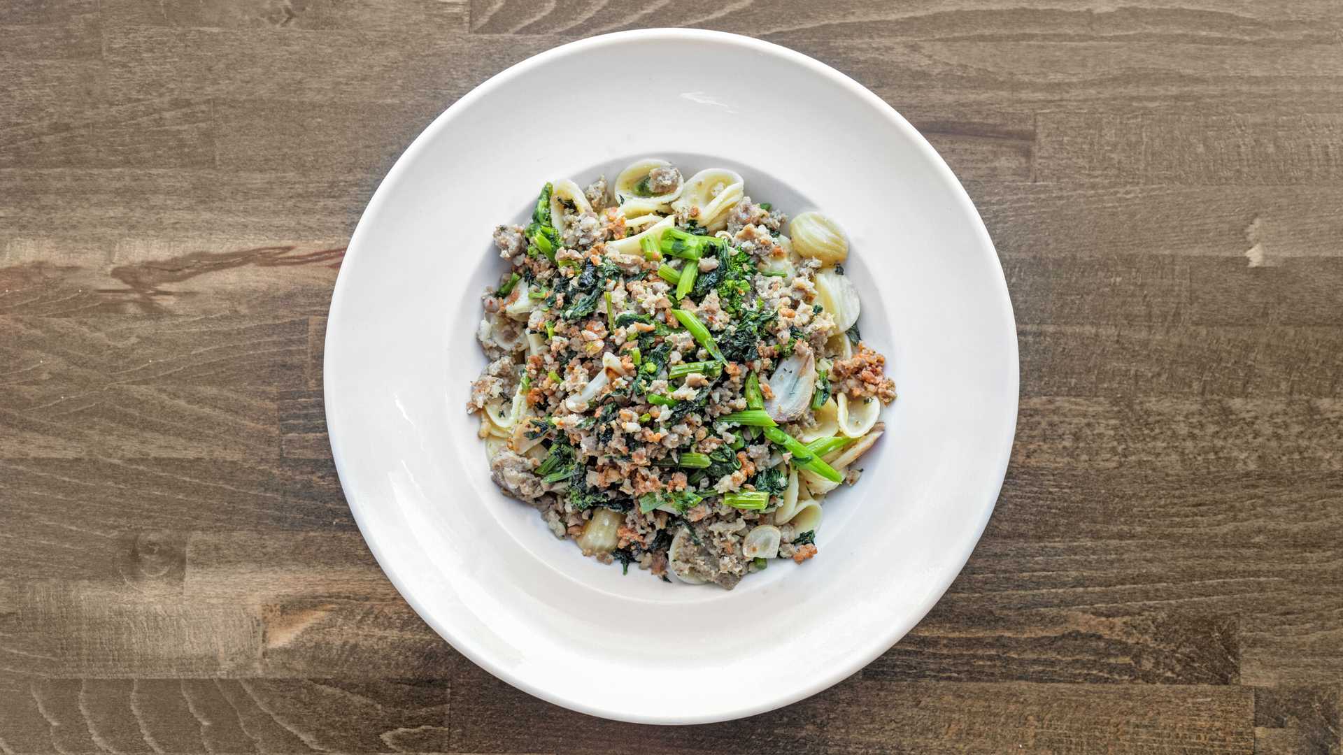 White plate with orecchiette pasta topped with sautéed ground meat, greens, and slices of garlic on a wooden table.