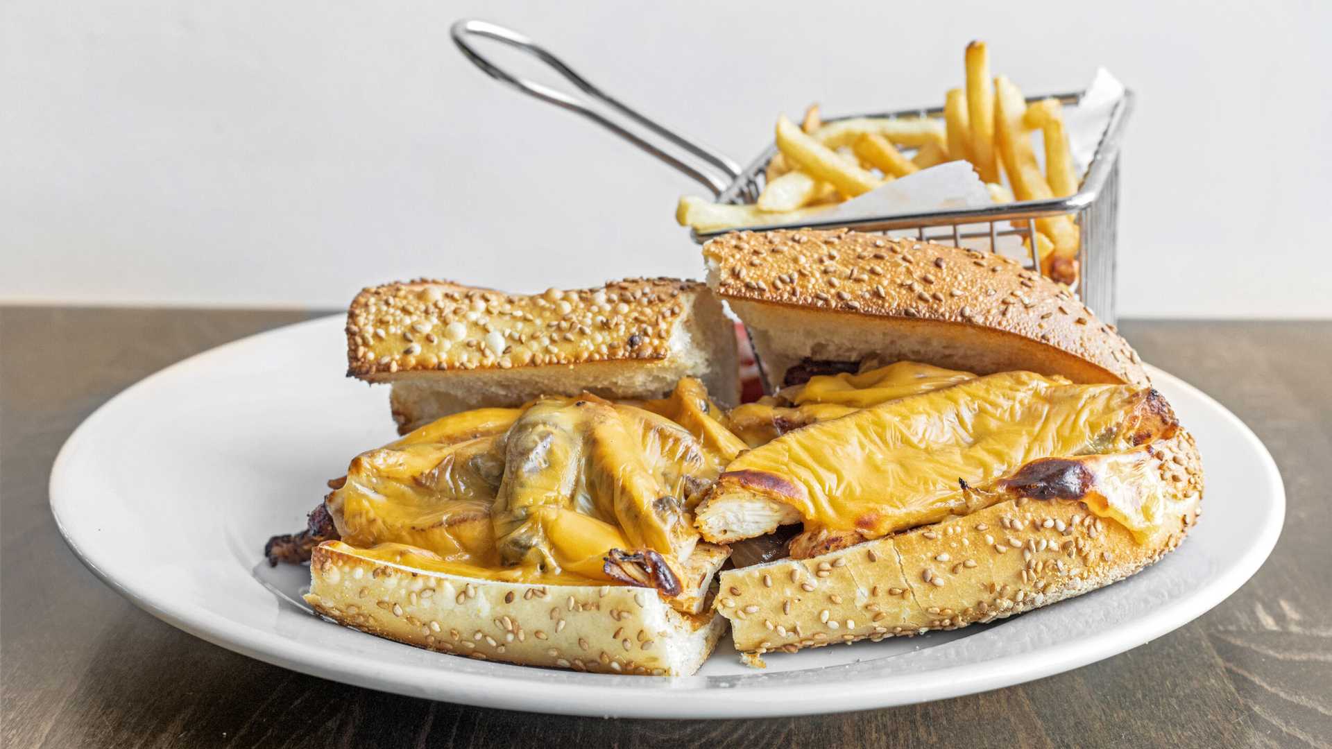 Grilled chicken Philly sandwich with melted cheese on sesame seed hoagie bun, served with fries in a metal basket.