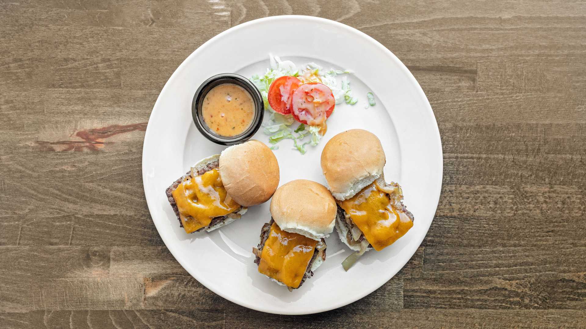 Three mini cheeseburgers with melted cheddar, onions, and a side of sauce, lettuce, and tomato on a white plate over a wooden table.