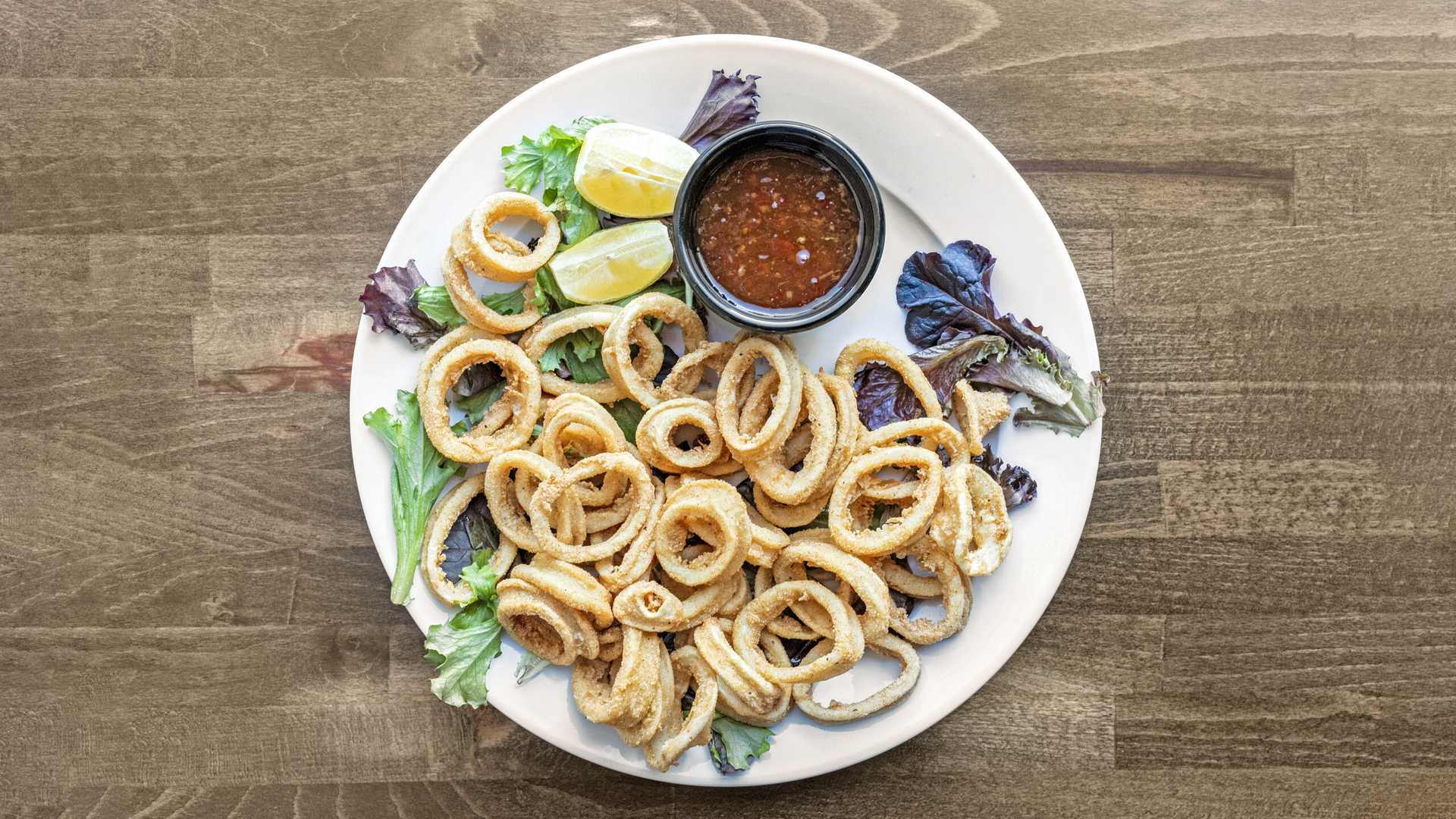 Plate of fried calamari rings served on mixed greens with lemon wedges and a cup of dipping sauce.