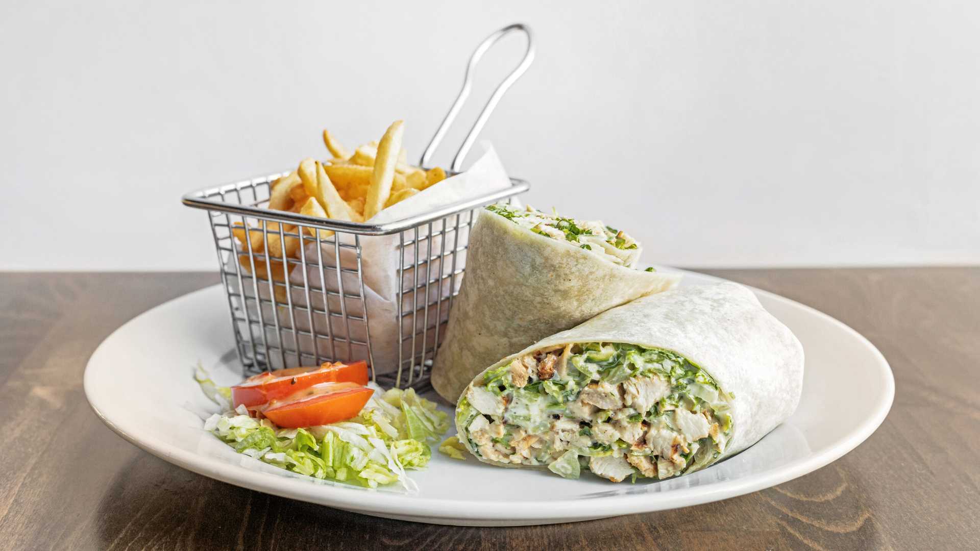 Grilled chicken Caesar wrap cut in half served with a small basket of French fries and a side of shredded lettuce and sliced tomato on a white plate.