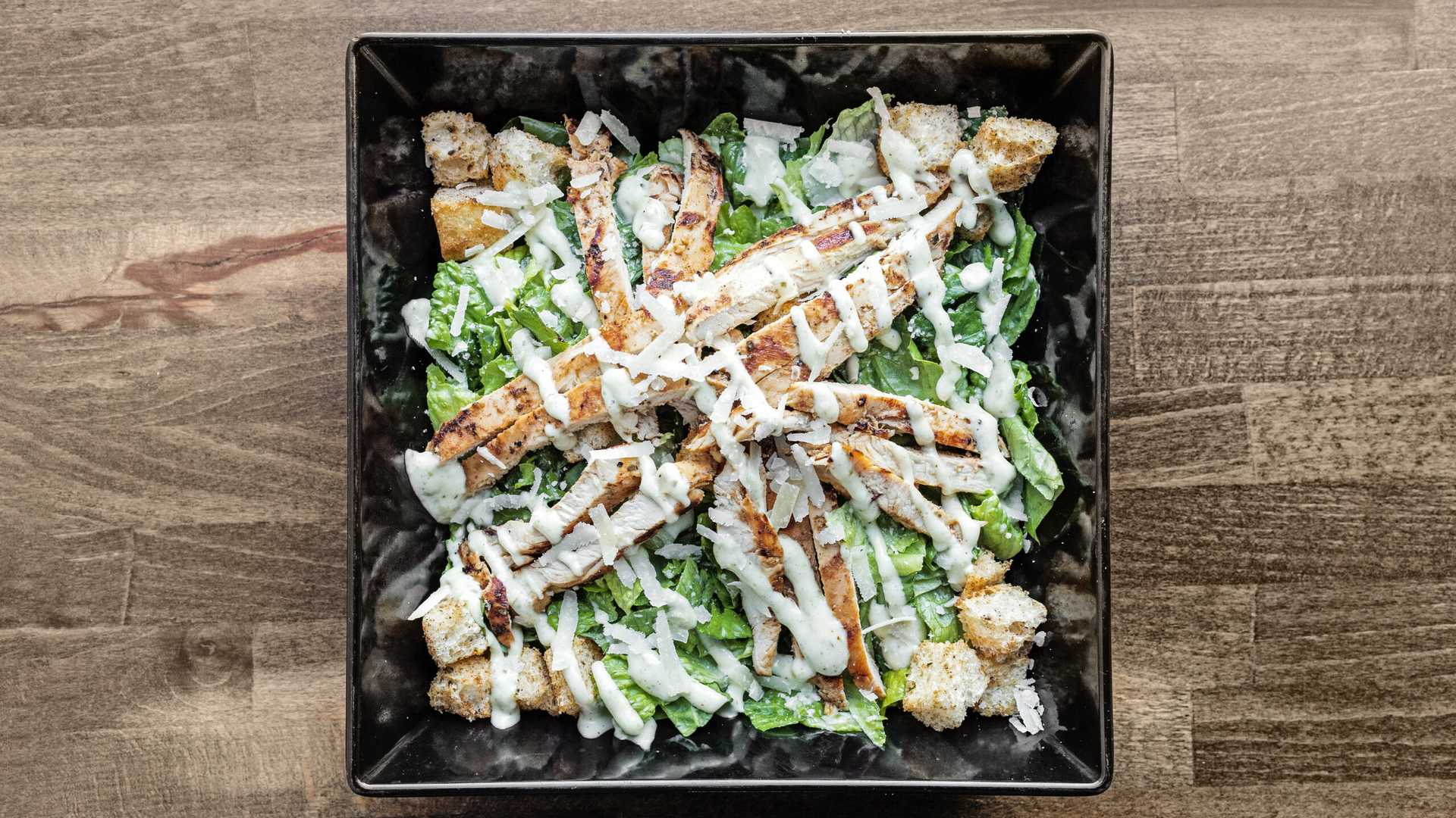 Grilled chicken Caesar salad with romaine lettuce, croutons, shaved Parmesan, and Caesar dressing in a black square bowl.
