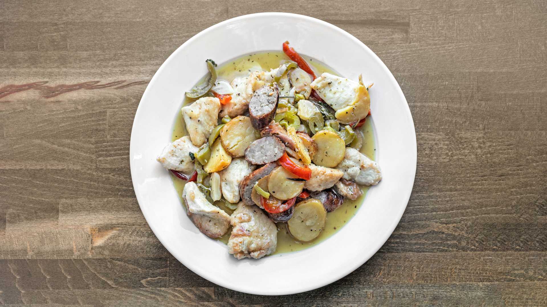 Plate of chicken scarpariello with sausage, roasted potatoes, and peppers in a light sauce on a wooden table.