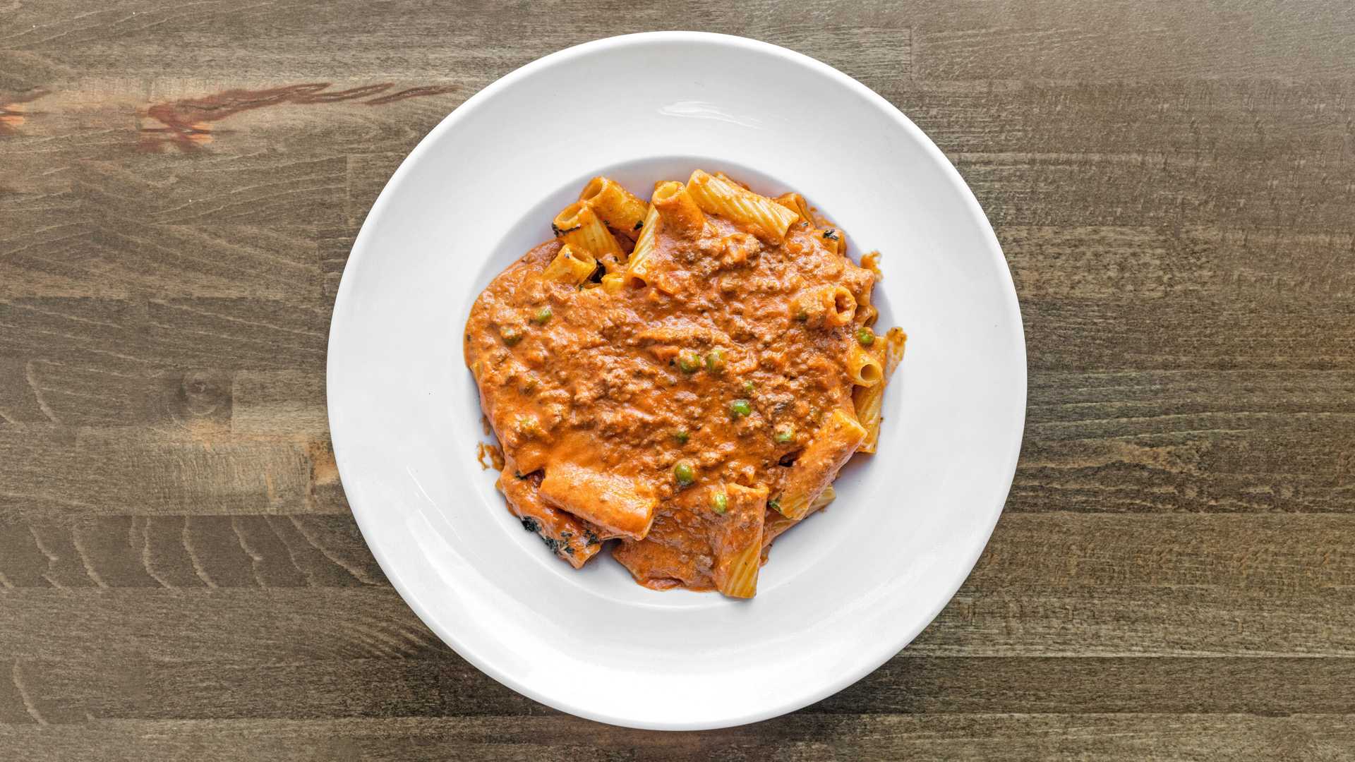 White plate with rigatoni pasta covered in thick meat sauce with peas, on a wooden surface.