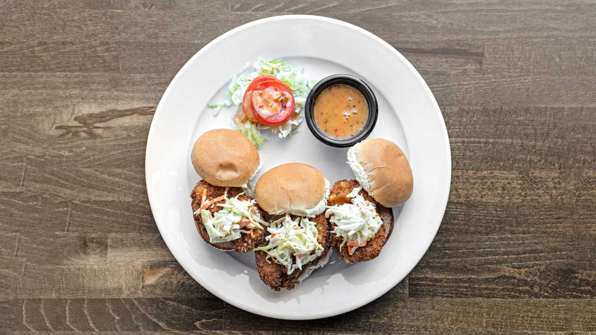 Three fried shrimp sliders topped with coleslaw on a plate with a small cup of sauce and a tomato slice on shredded lettuce.